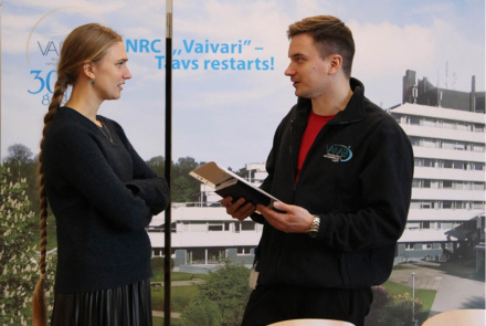 Sieviete un vīrietis stāv iekštelpās un sarunājas; vīrieša rokās ir piezīmju grāmatiņa un pildspalva. Aiz viņiem ir liels plakāts ar ēkas attēlu un uzrakstu "NRC "Vaivari" - Tavs atsāk darbu!".