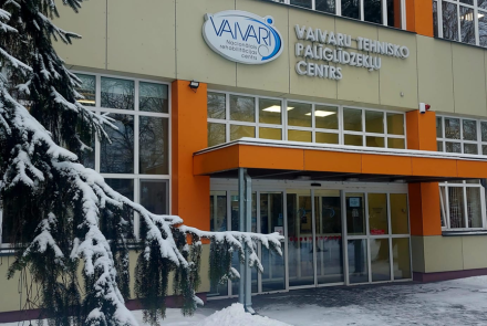 Ieeja ēkā ar lieliem logiem un oranžu markīzi, uz kuras ir uzraksts VAIVARI Tehnisko Palīgierīču Centrs. Sniegs klāj zemi un koku zarus ēkas priekšā.