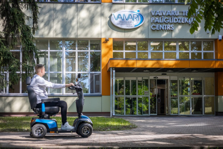 Vīrietis brauc ar zilu skūteri pa ietvi pie ēkas ar uzrakstu Vaivaru Palīglīdzekļu centrs ar lieliem logiem un izkārtni virs ieejas.
