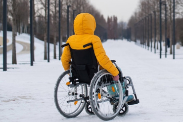 Cilvēks dzeltenā žaketē sēž ratiņkrēslā uz sniegota celiņa, kas aukstā, drēgnā un drēgnā dienā izklāts ar kokiem un laternu stabiem.