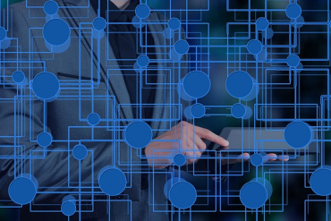 Persona uzvalkā izmanto planšetdatoru, uz kura ir zilas, savstarpēji savienotas ķēdes veida līnijas un mezgli, kas simbolizē tehnoloģiju un digitālos tīklus.