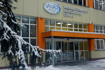 Ieeja ēkā ar lieliem logiem un oranžu markīzi, uz kuras ir uzraksts VAIVARI Tehnisko Palīgierīču Centrs. Sniegs klāj zemi un koku zarus ēkas priekšā.