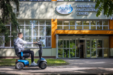 Vīrietis brauc ar zilu skūteri pa ietvi pie ēkas ar uzrakstu Vaivaru Palīglīdzekļu centrs ar lieliem logiem un izkārtni virs ieejas.