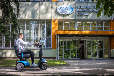 Vīrietis ar zilu skrejriteni brauc pa celiņu pie Vaivaru Nacionālā rehabilitācijas centra ēkas, kurai ir lieli logi, koki un izkārtne virs ieejas.