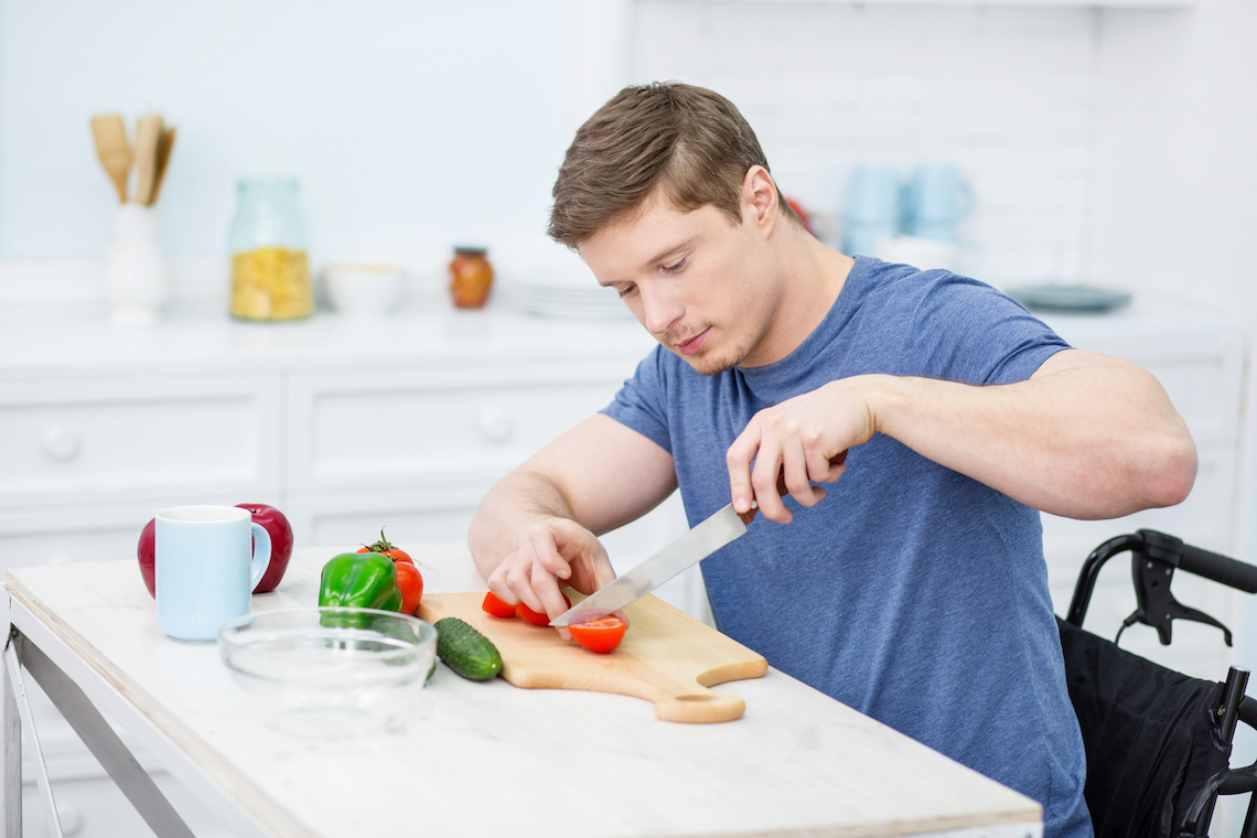 Vīrietis ratiņkrēslā sagriež tomātu uz koka dēlīša gaišā virtuvē. Uz galda ir gurķis, paprika, krūzes un bļoda. Viņš ir koncentrējies uz ēdiena gatavošanu.